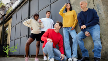 A lively group of young adults hanging out in front of a large window, each wearing a different colored SU7600 crewneck sweatshirt by Supasoft Apparel, with a Awkward Styles logo in the corner.