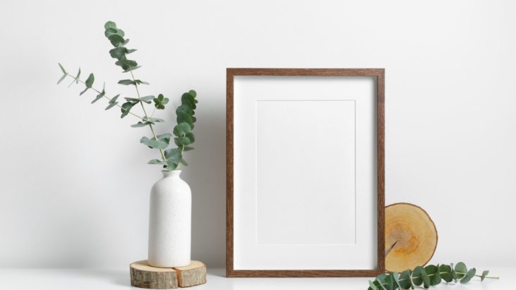 blank framed canvas on top of a white table in front of a white background and next to a white vase with a plant inside