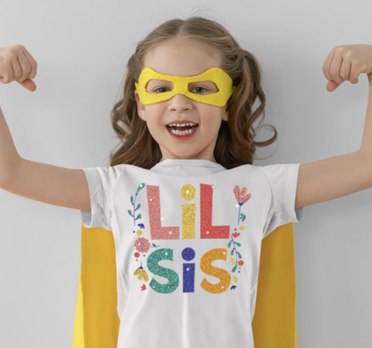 Girl with a yellow superhero cape and yellow eye mask wearing a white tshirt that says "lil sis"
